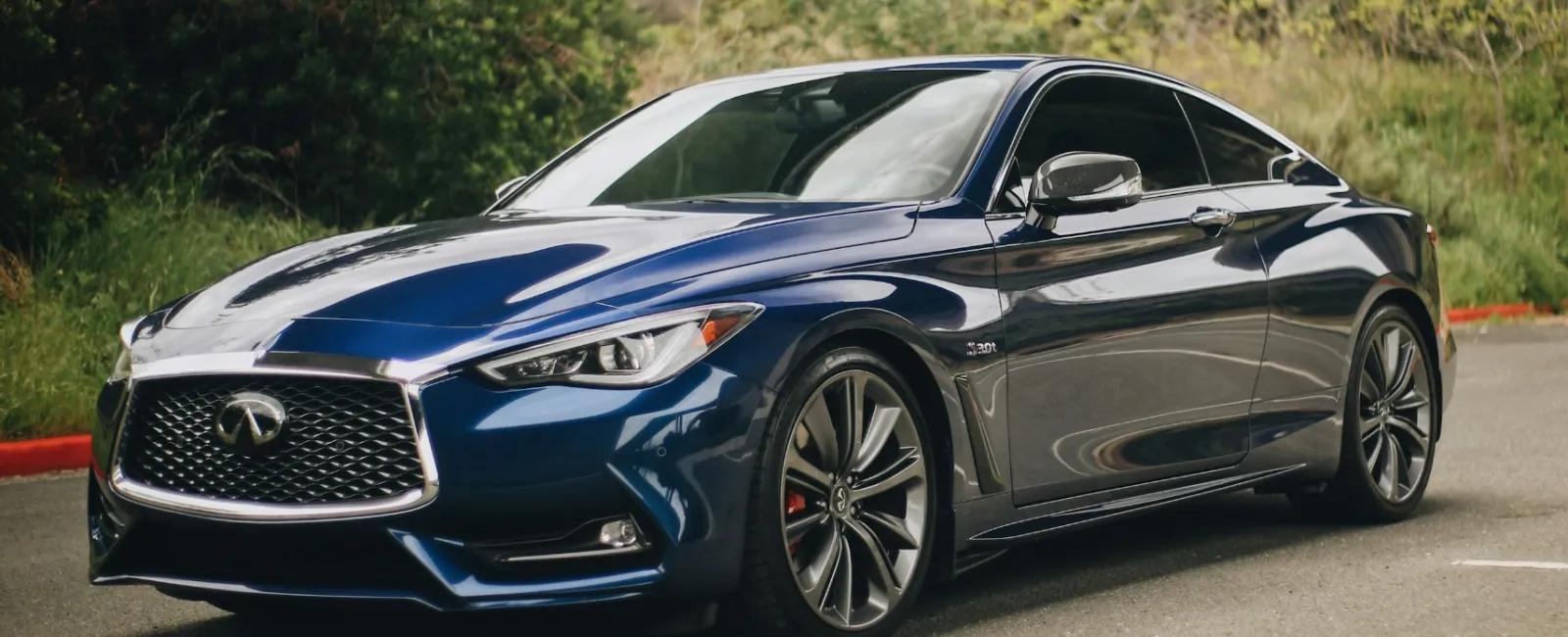 Blue Infiniti sports sedan parked outdoors near greenery and residential buildings under an overpass.