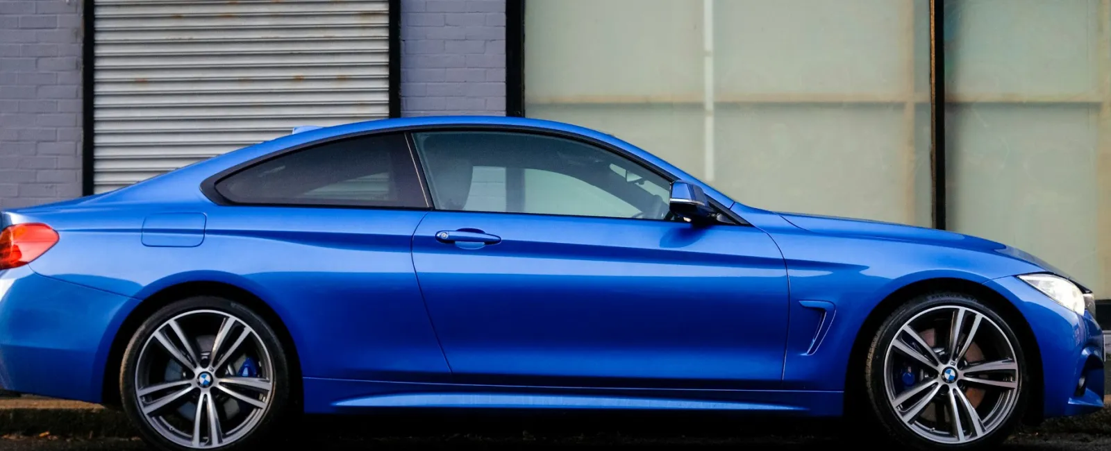 Sleek blue BMW coupe parked on street in front of gray brick building with 'Entrance' sign above door.