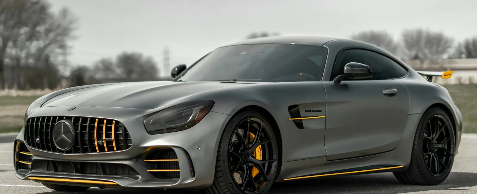 Sleek gray Mercedes AMG GT sports car with black and yellow accents parked on a road with blurred trees in background