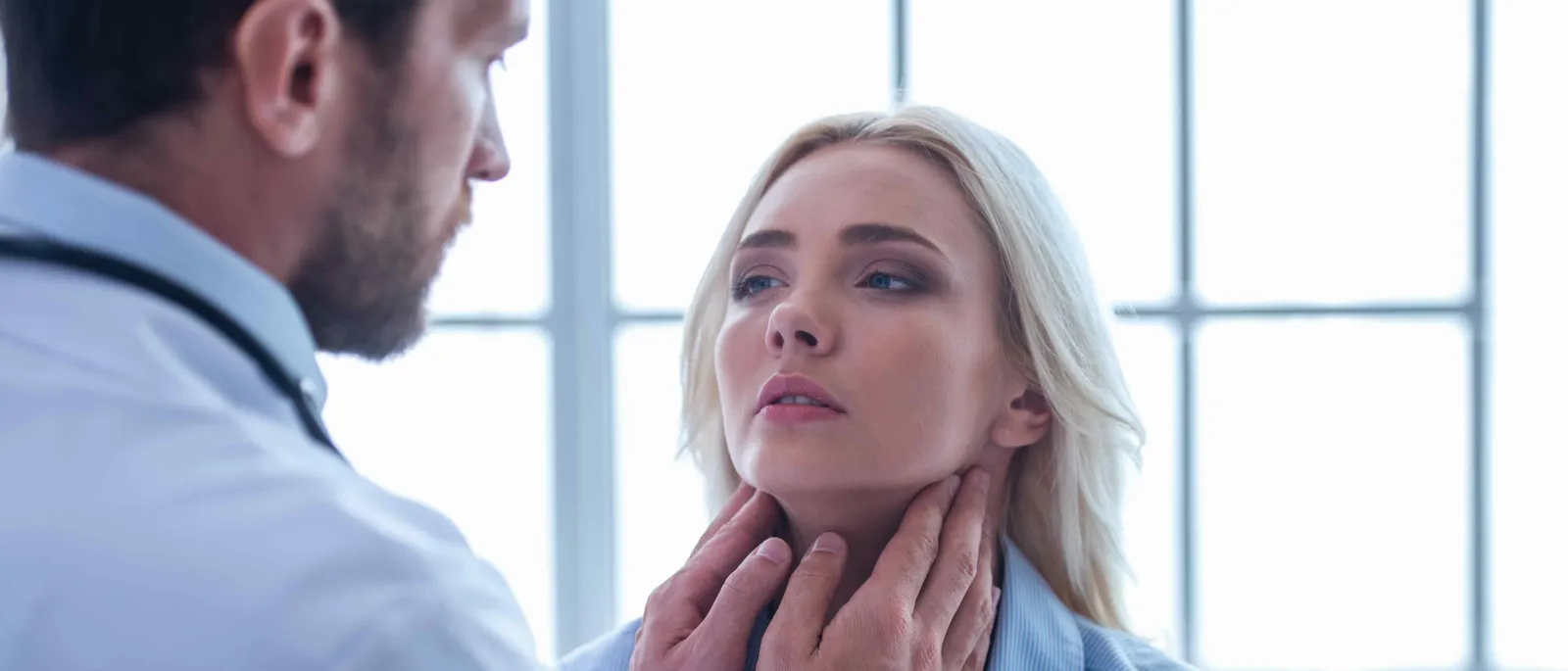 Doctor examining woman's throat in medical office with large windows and bright natural light.