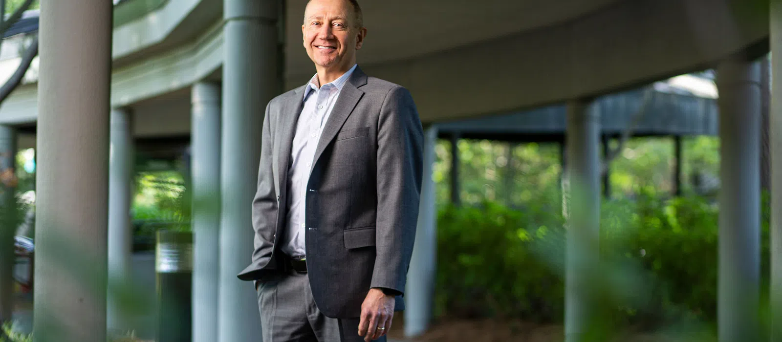 Smiling businessman in gray suit standing outdoors near curved modern architectural columns with greenery