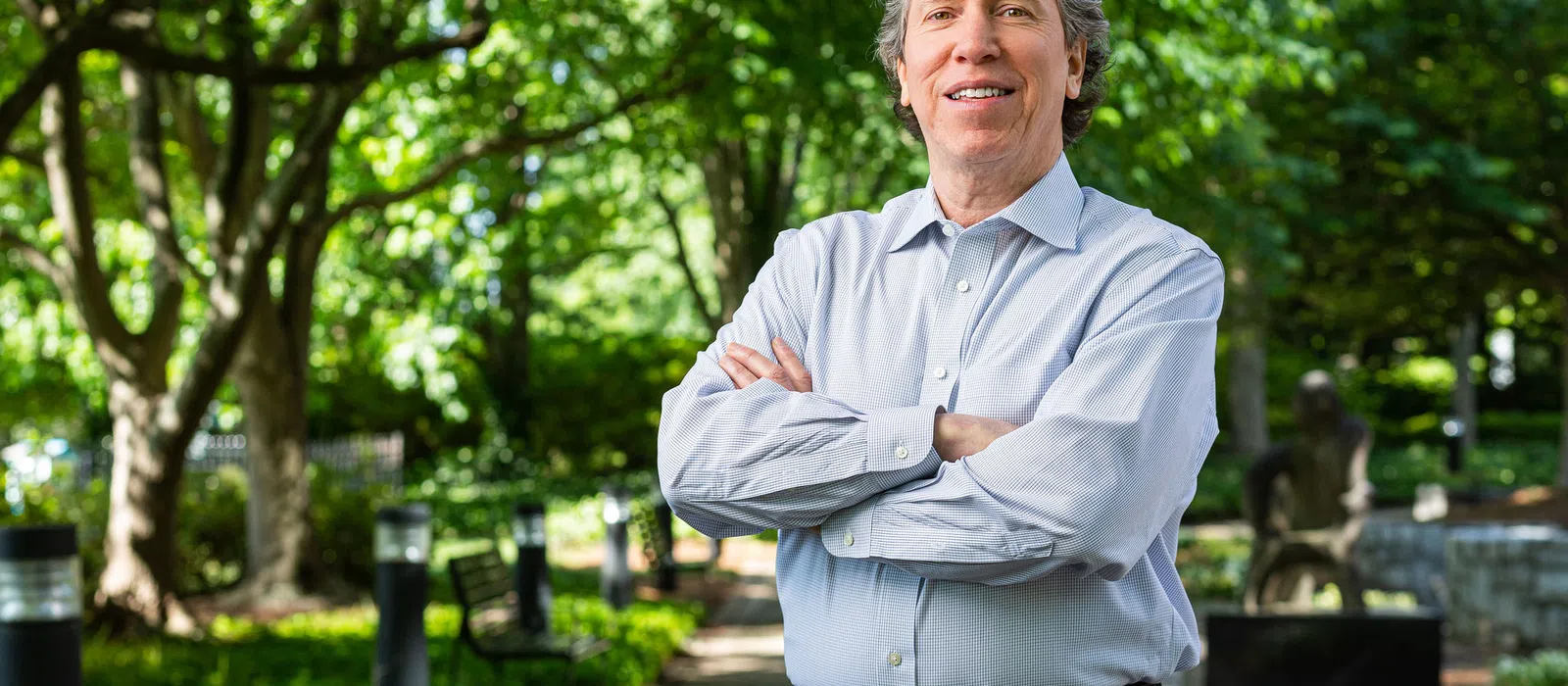 Smiling man with gray hair in light blue shirt standing with arms crossed on a sunny park pathway with trees.