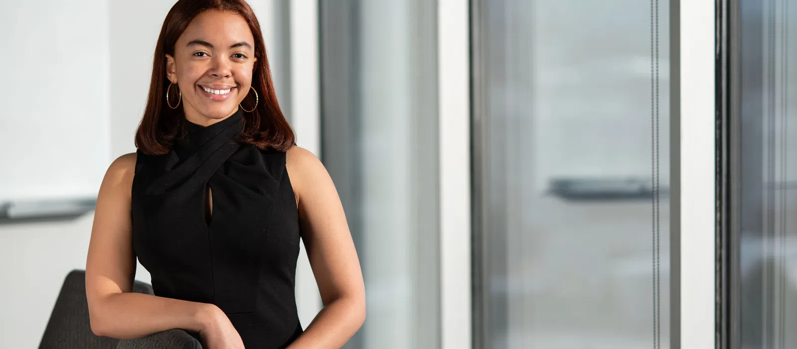 Smiling woman in black dress standing confidently in a modern office with large windows and chairs.