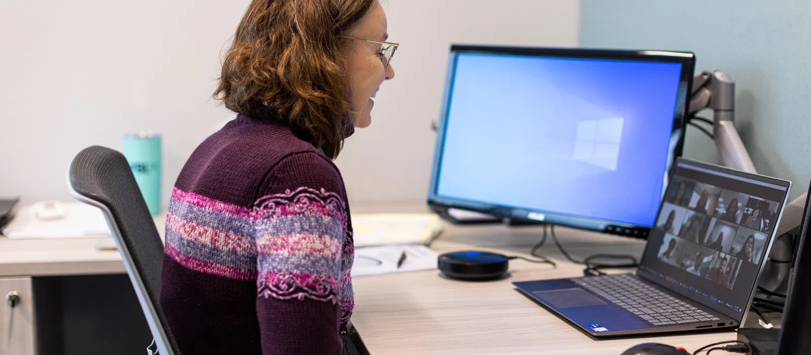 Woman in purple sweater participates in a video conference at her office desk with dual computer monitors.