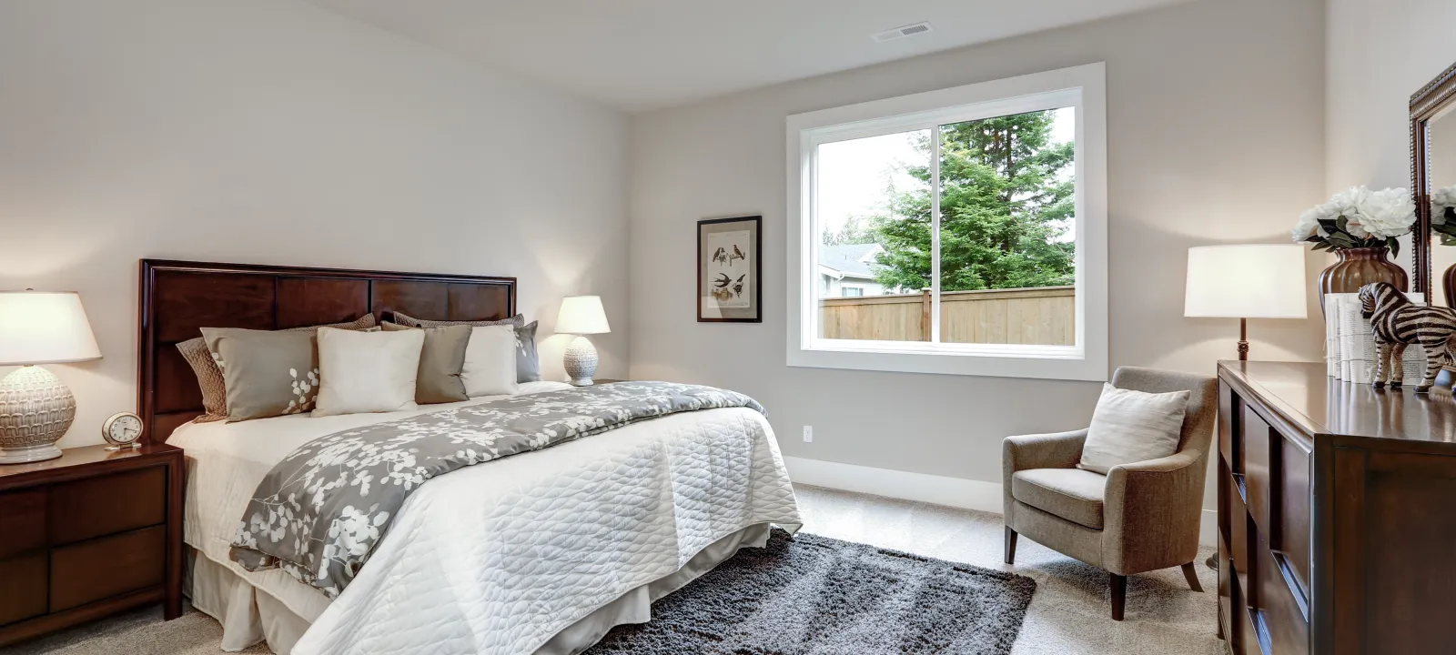 Bright bedroom with wooden furniture, queen bed, gray rug, armchair, and large window overlooking greenery.