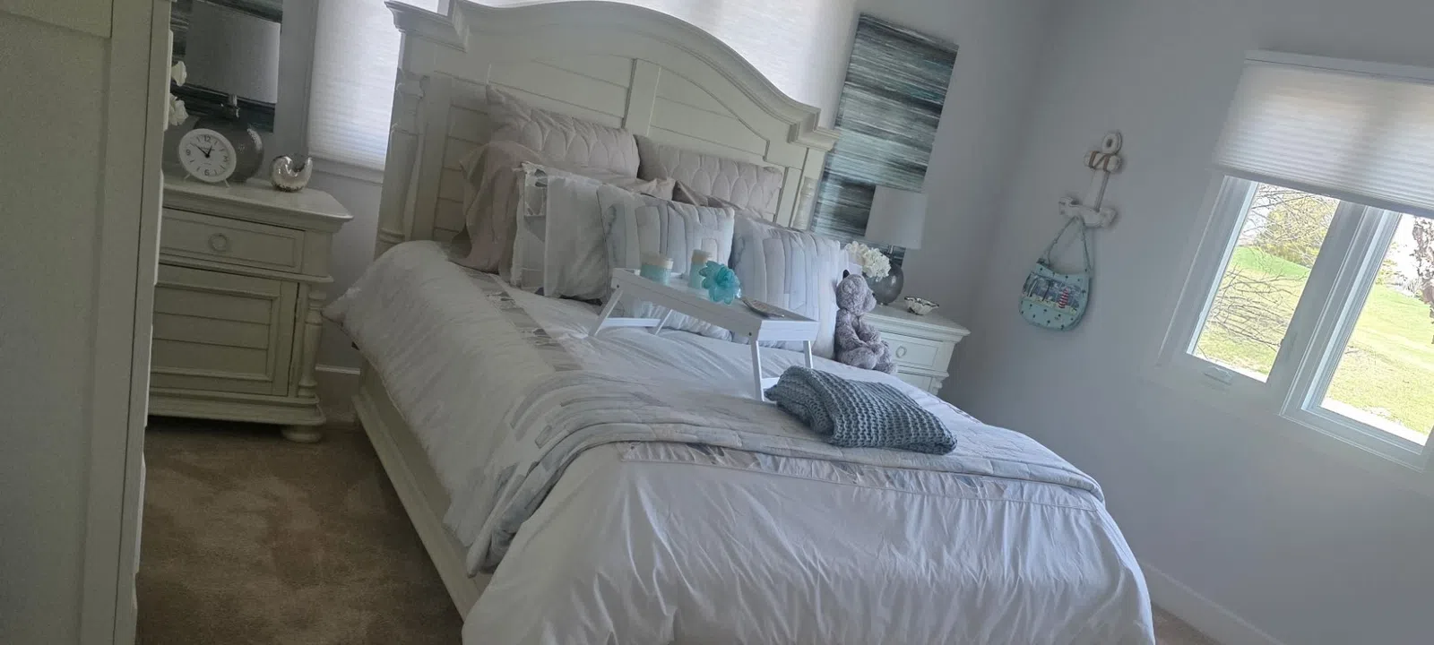 Bright bedroom with white wooden furniture, plush carpet, large windows, and neatly made bed with decorative pillows.