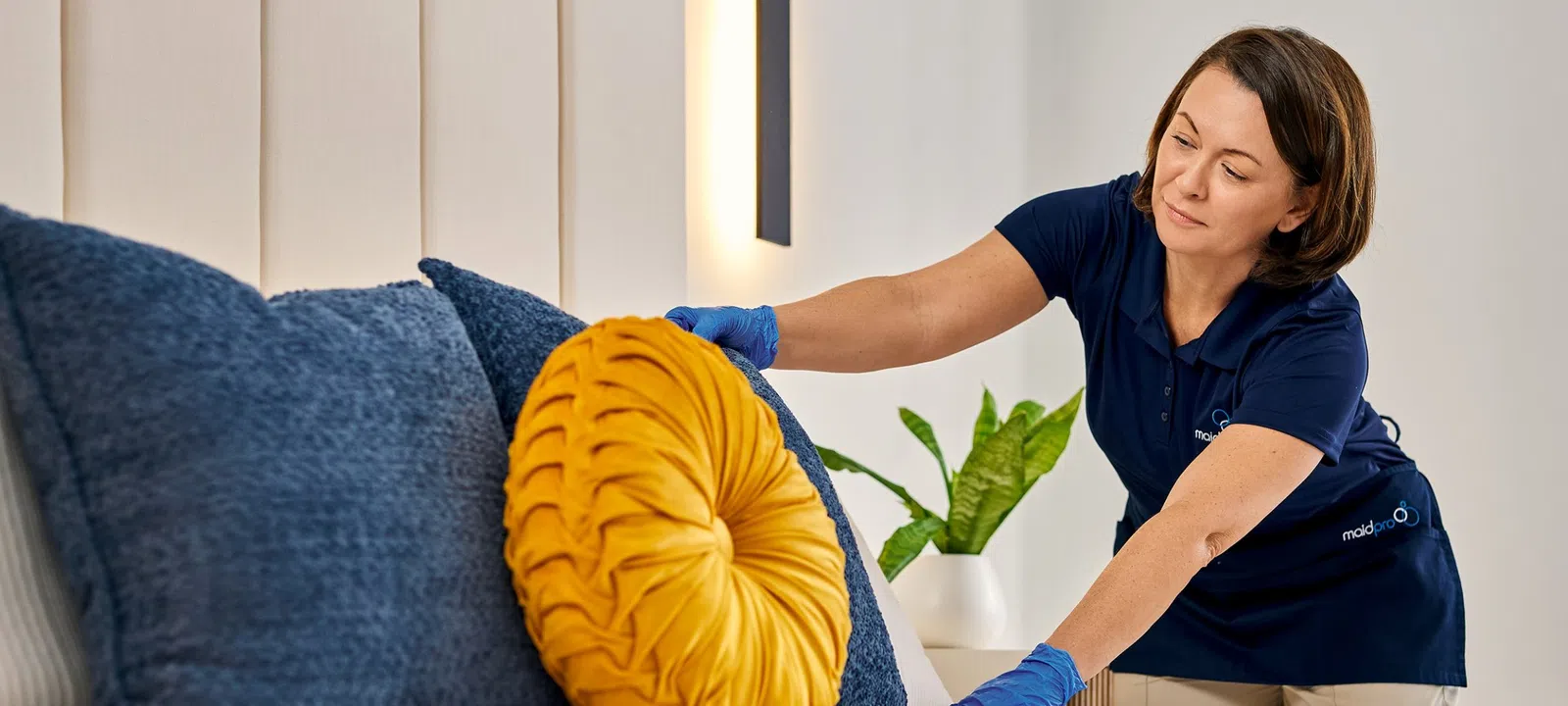 House cleaner arranging decorative pillows on a bed in a modern bedroom with beige and blue tones.