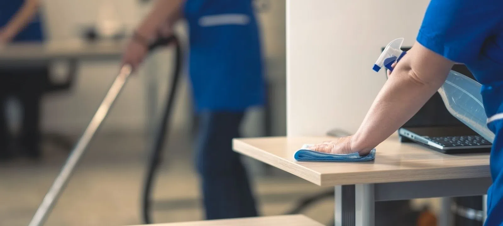 Professional cleaners in blue uniforms wiping desk and vacuuming office floor for sanitation.