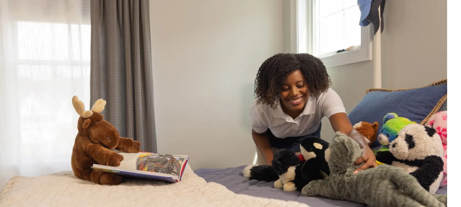 Smiling woman arranging stuffed animals on bed with moose toy reading a book nearby in cozy bedroom.