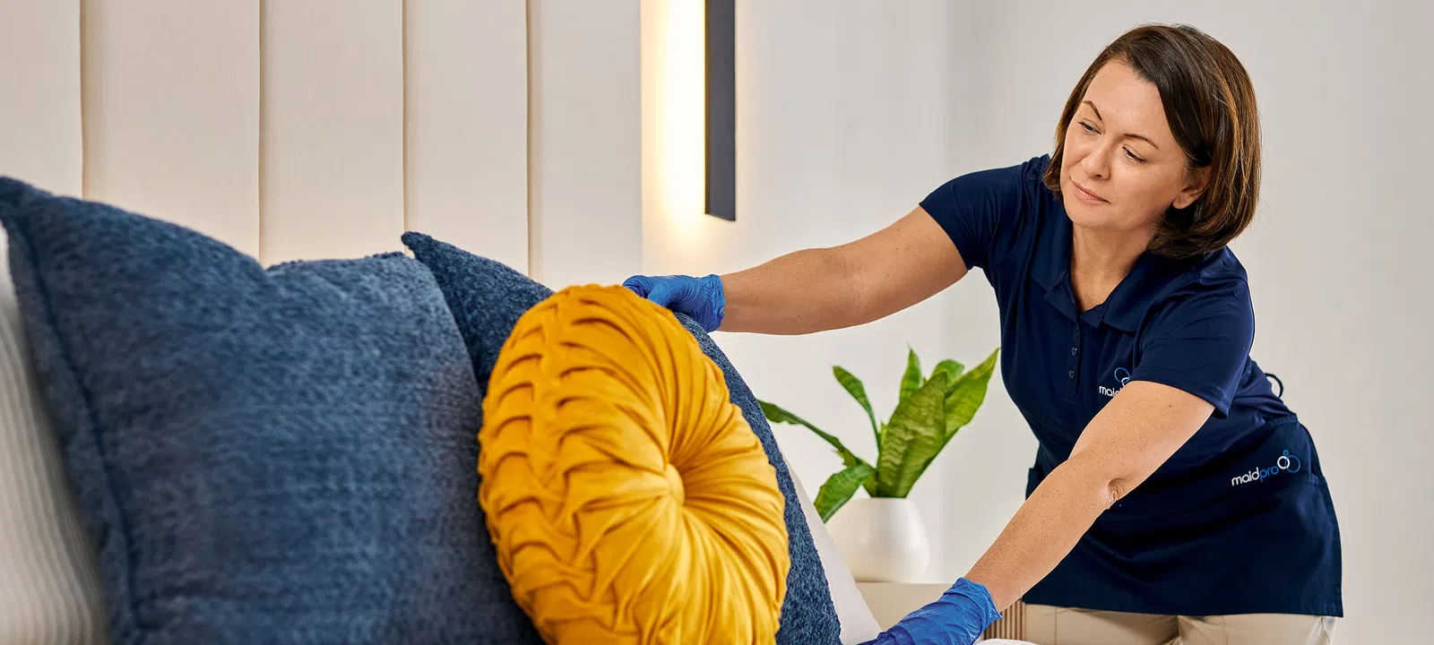 Housekeeper arranging yellow round pillow on bed with blue cushions in bright modern bedroom.