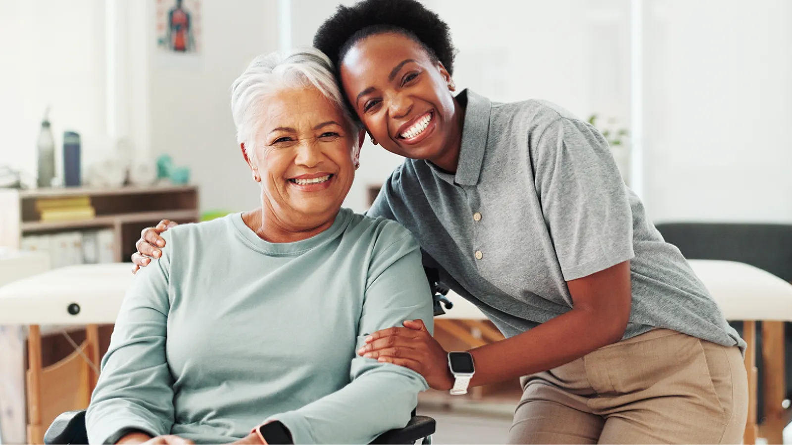 Smiling elderly woman in wheelchair embraced by friendly caregiver in bright living room setting