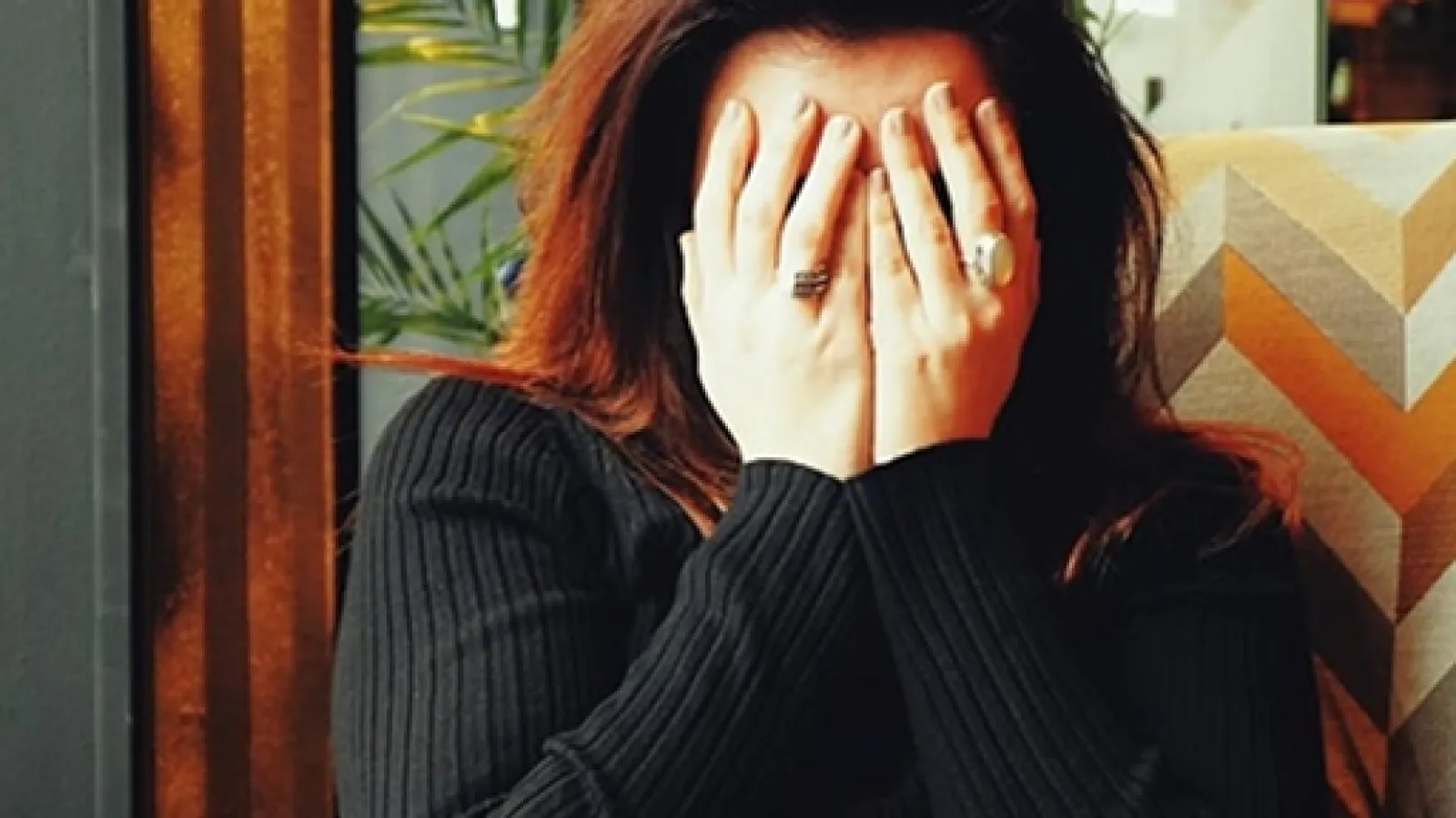 Woman in black sweater covering her face with hands sitting indoors near a plant and patterned chair.