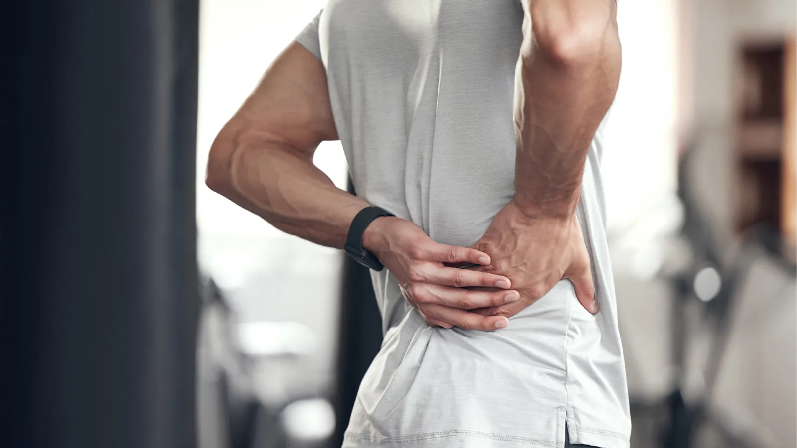 Man in gray shirt holding lower back in pain, indicating muscle strain or injury during workout.