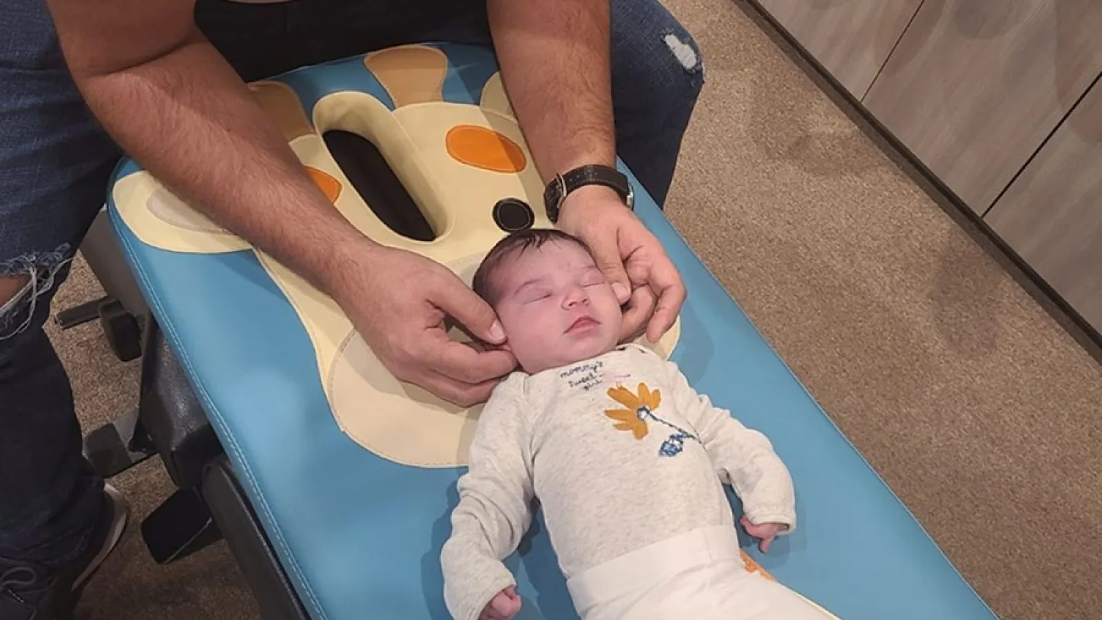 Man gently holding a baby's head while baby lies on a blue giraffe-themed table in a cozy room.