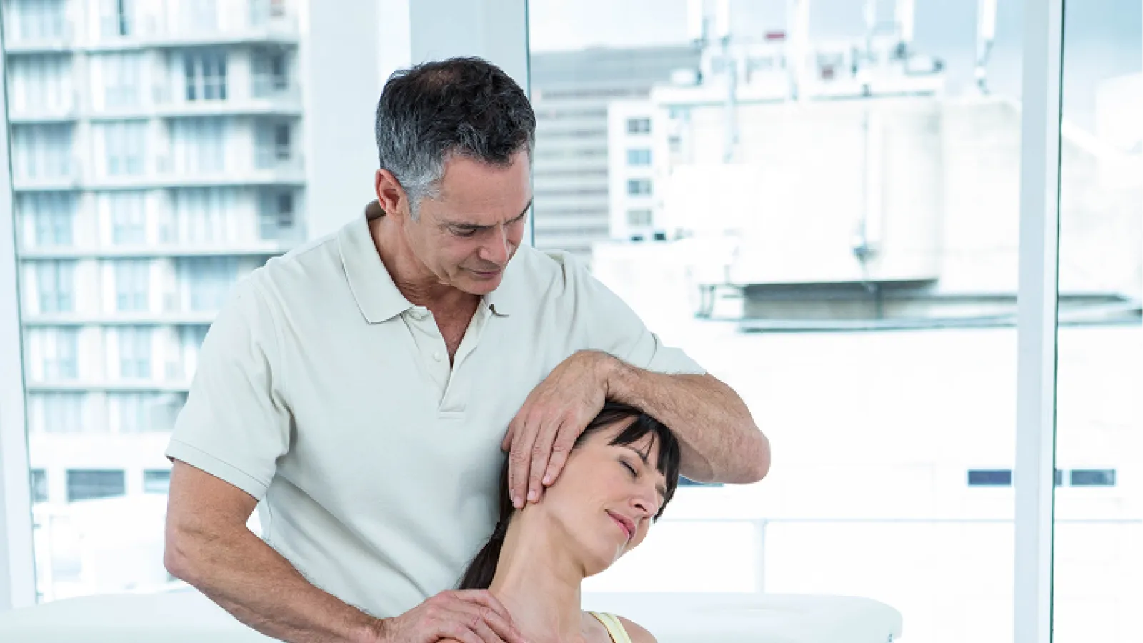 Pregnant woman on exercise ball receiving neck stretch from male physiotherapist in bright therapy room.