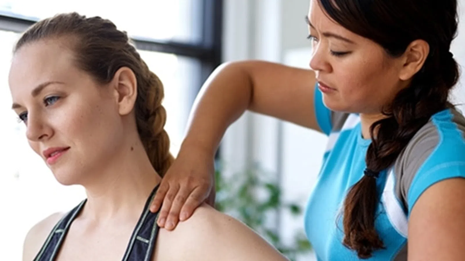 Female physiotherapist assisting woman with shoulder stretch in bright clinic setting.