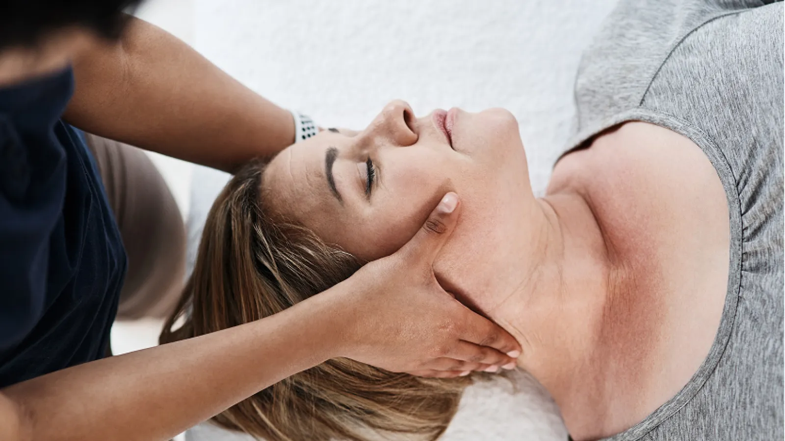 Woman receiving relaxing facial massage therapy lying down with eyes closed wearing gray shirt