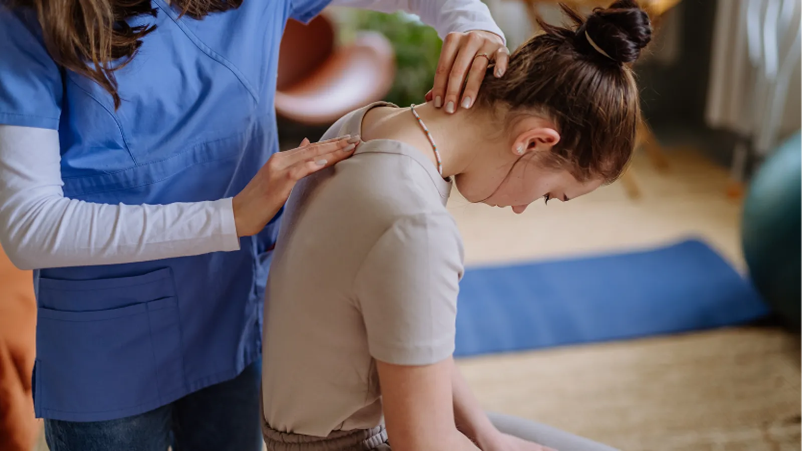 Physical therapist assisting a woman with shoulder stretch in a bright therapy room with large windows.