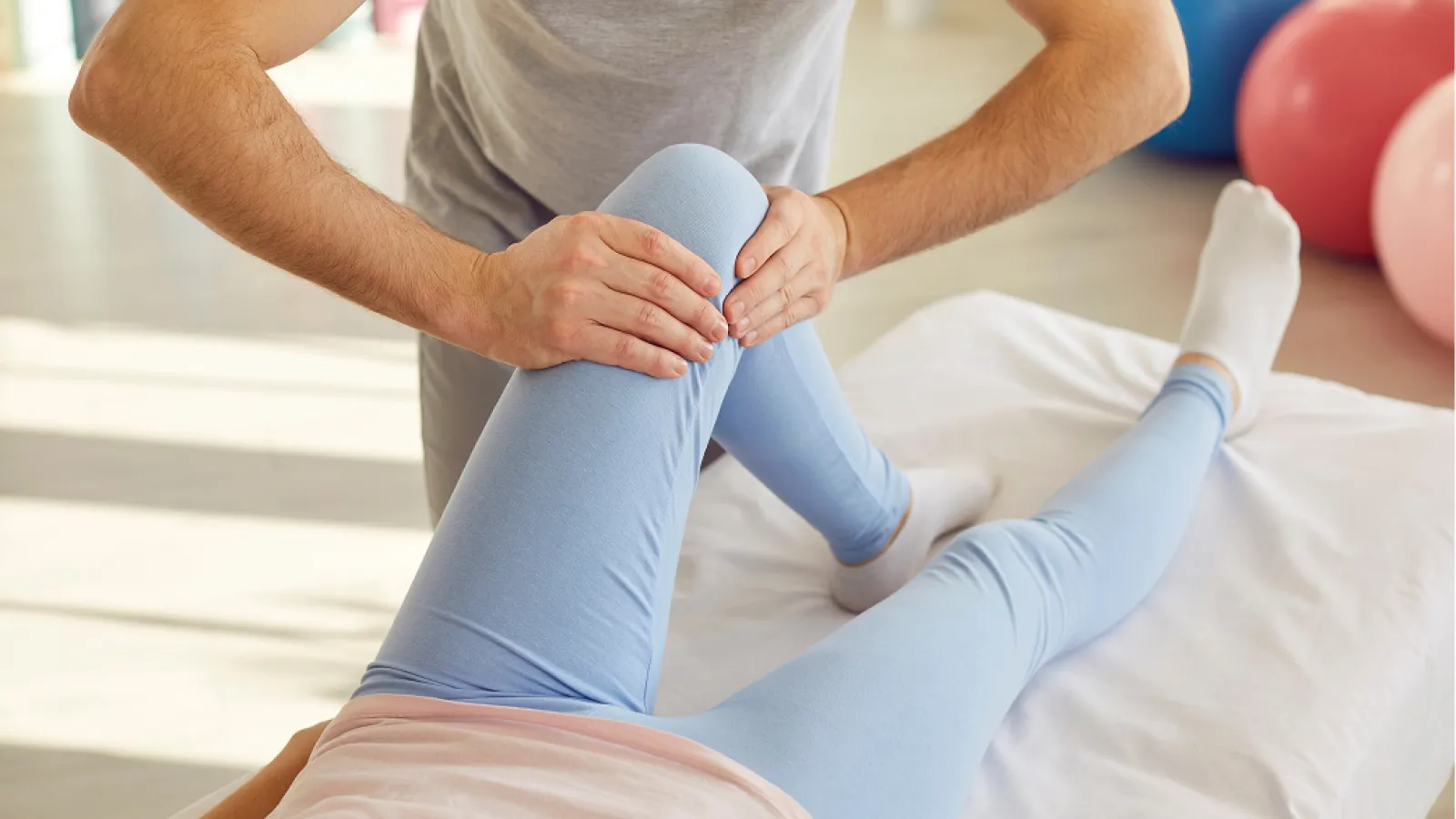 Physical therapist adjusting patient's bent knee during rehabilitation session indoors with exercise balls nearby.