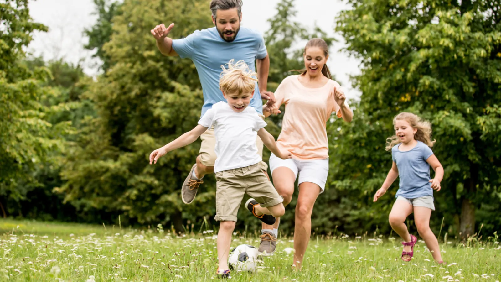 Family of four happily playing soccer together outdoors on a green field with trees in the background