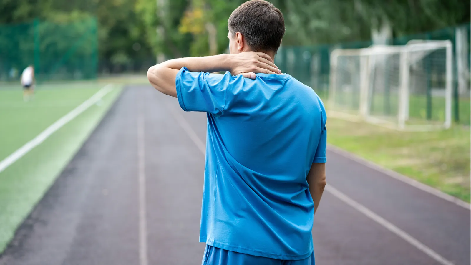 Man in blue sportswear holding his neck in pain standing on a running track near a soccer field.