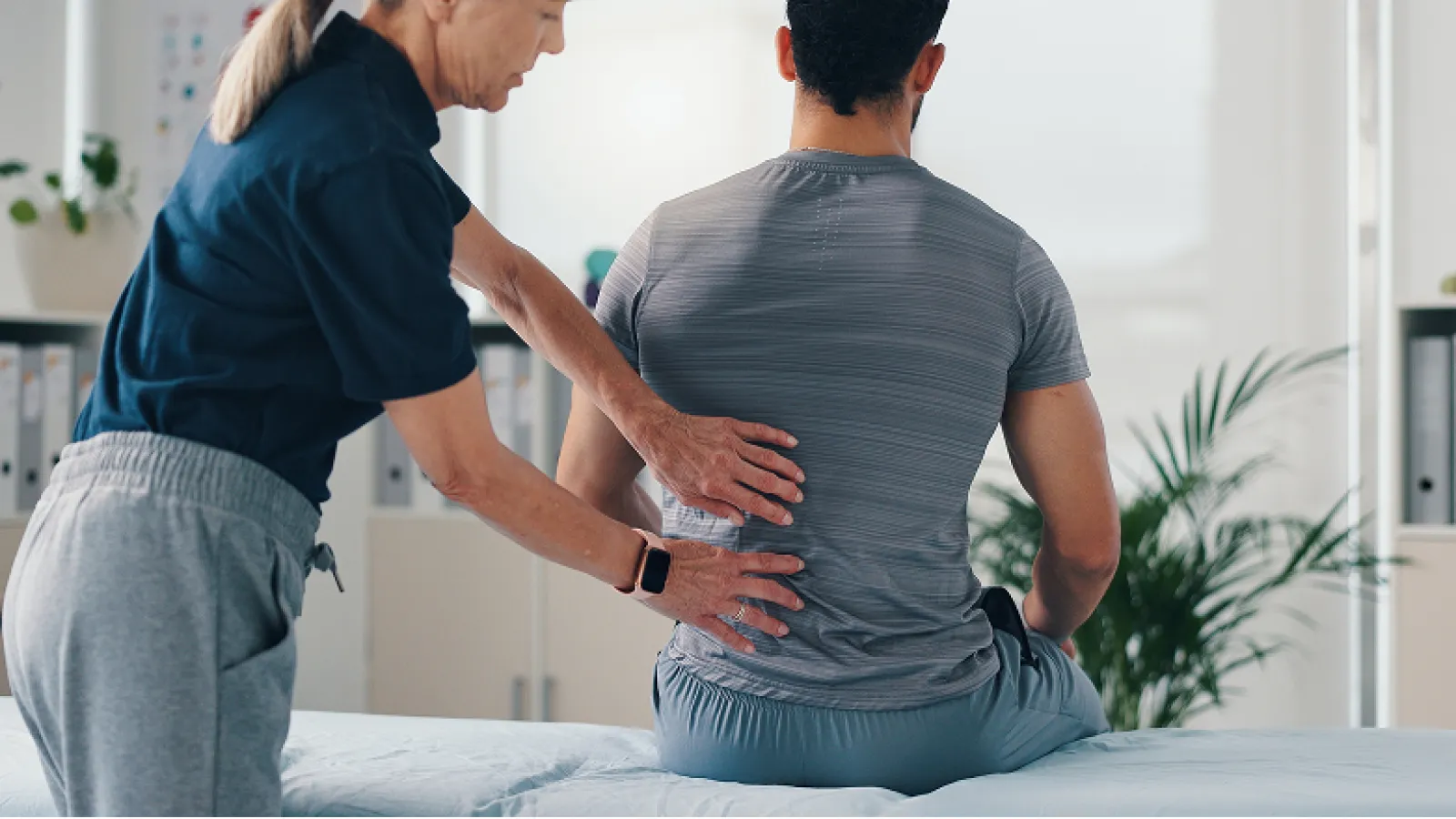 Physical therapist examining a man's lower back in a bright clinic setting with plants and shelves.