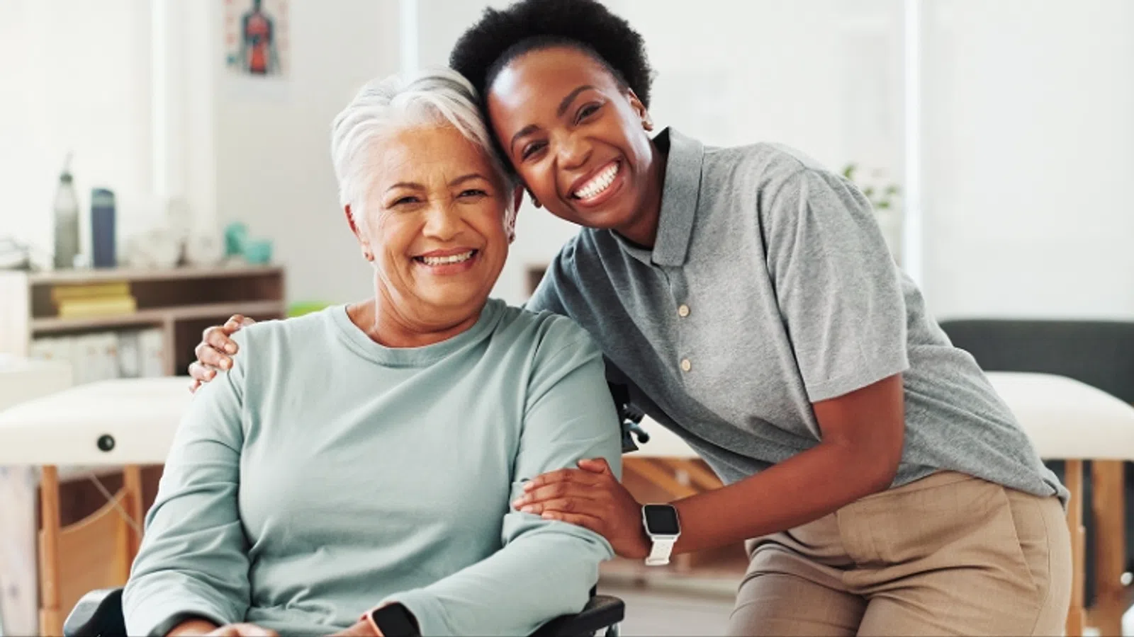 Smiling elderly woman in wheelchair with a caring young woman hugging her in a bright room.