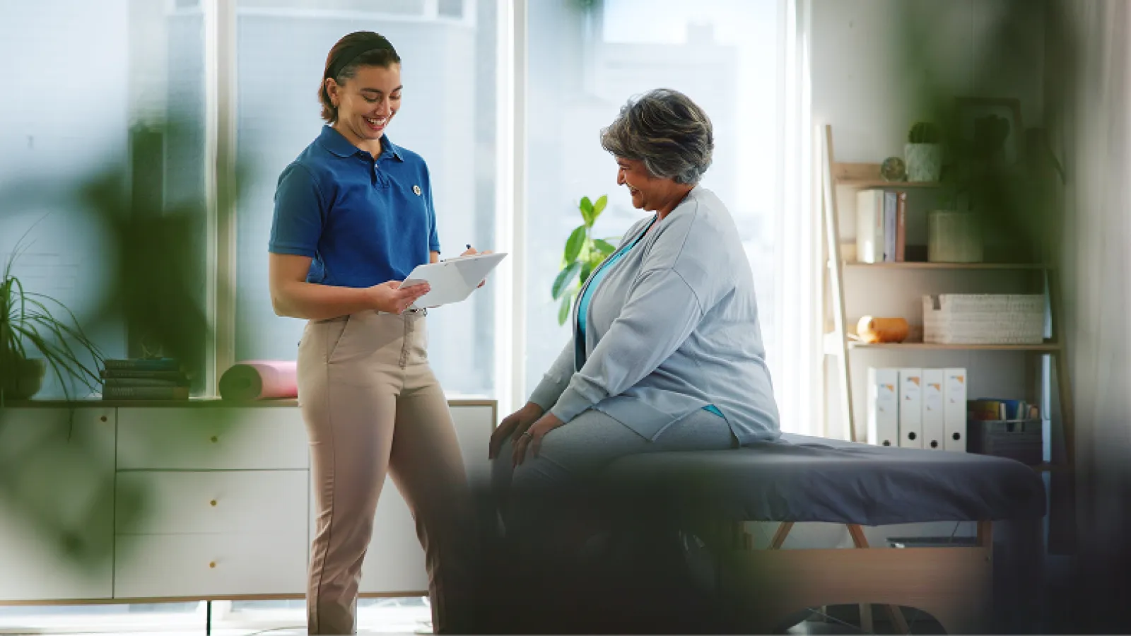 Therapist discussing notes with a smiling senior woman sitting on a medical examination table in a bright room