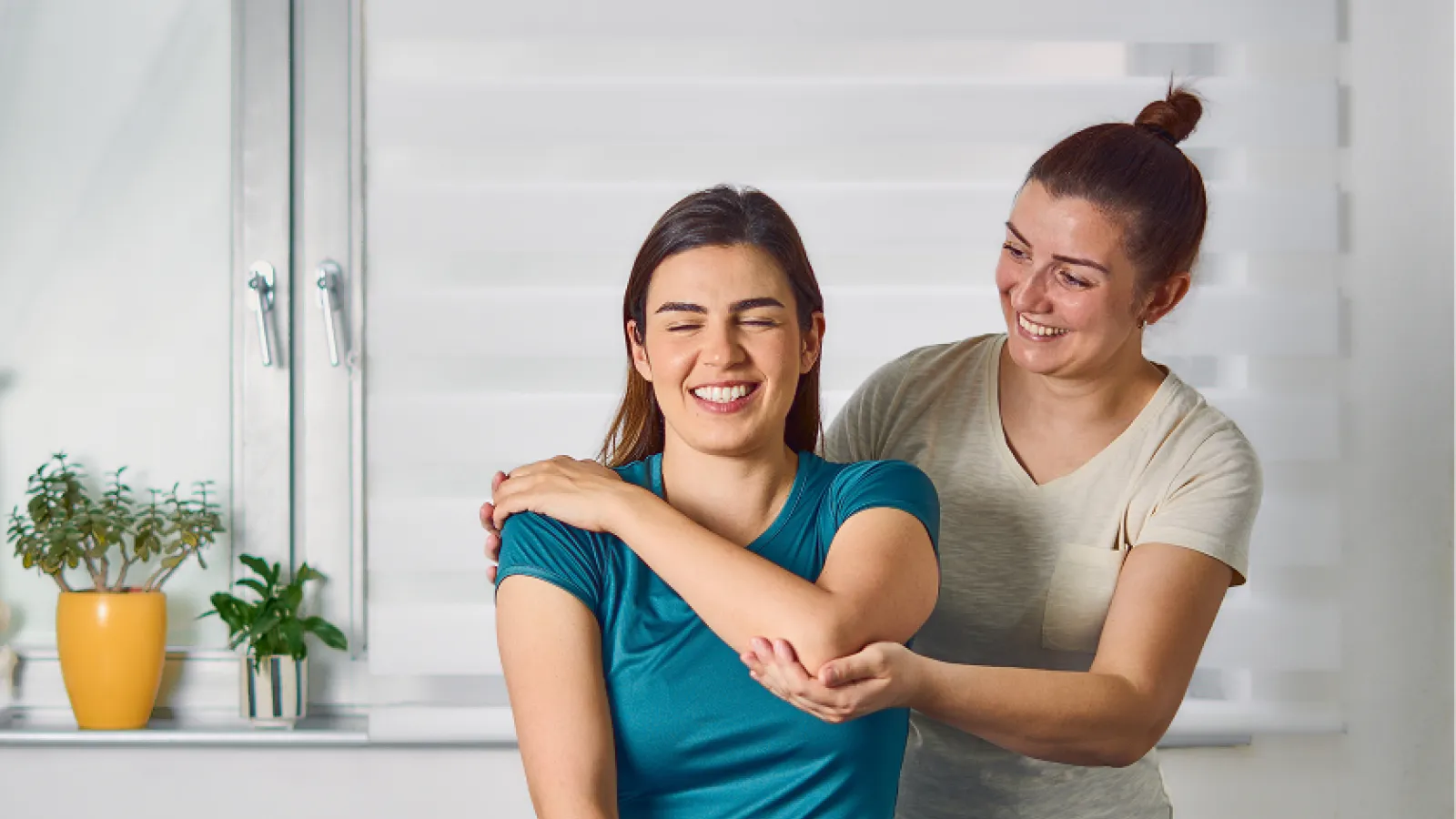 Physical therapist assisting young woman in shoulder stretch exercise in a bright room with plants.
