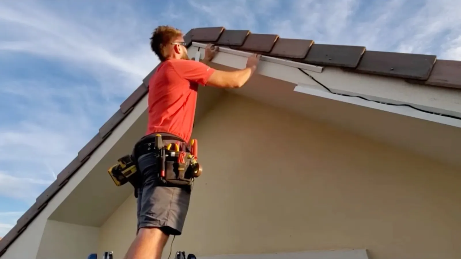Man in red shirt installing or repairing roof tiles on a house under a blue sky.