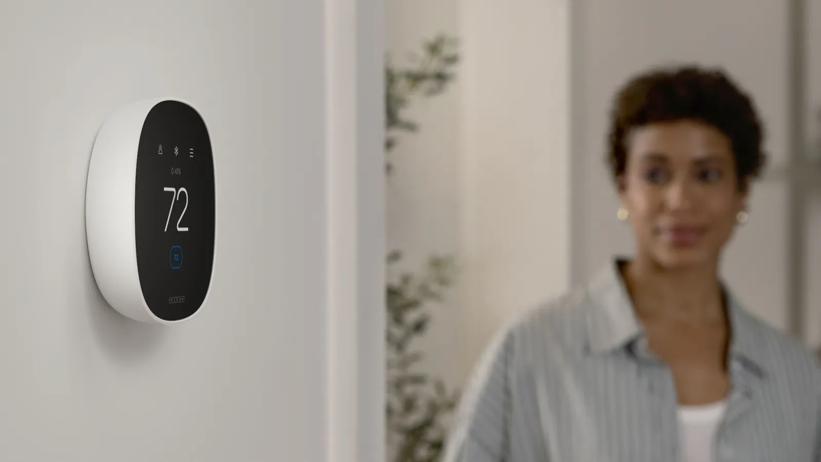 Modern smart thermostat set to 72 degrees mounted on wall with woman in the background