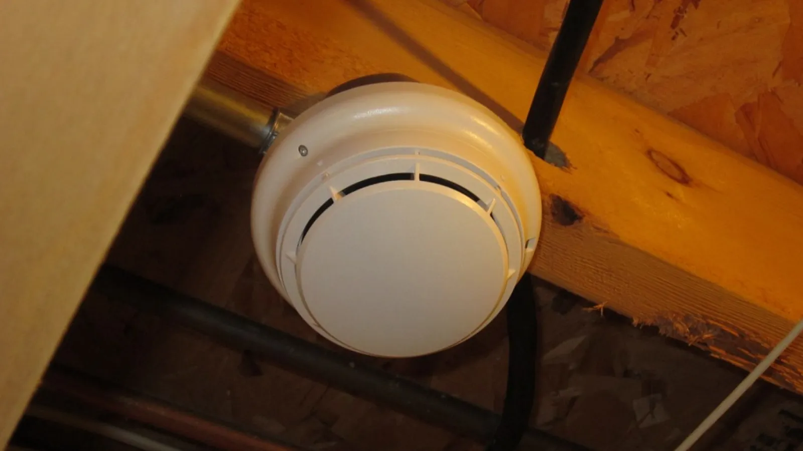 Round white smoke detector mounted on wooden ceiling beams in attic space with exposed wiring.