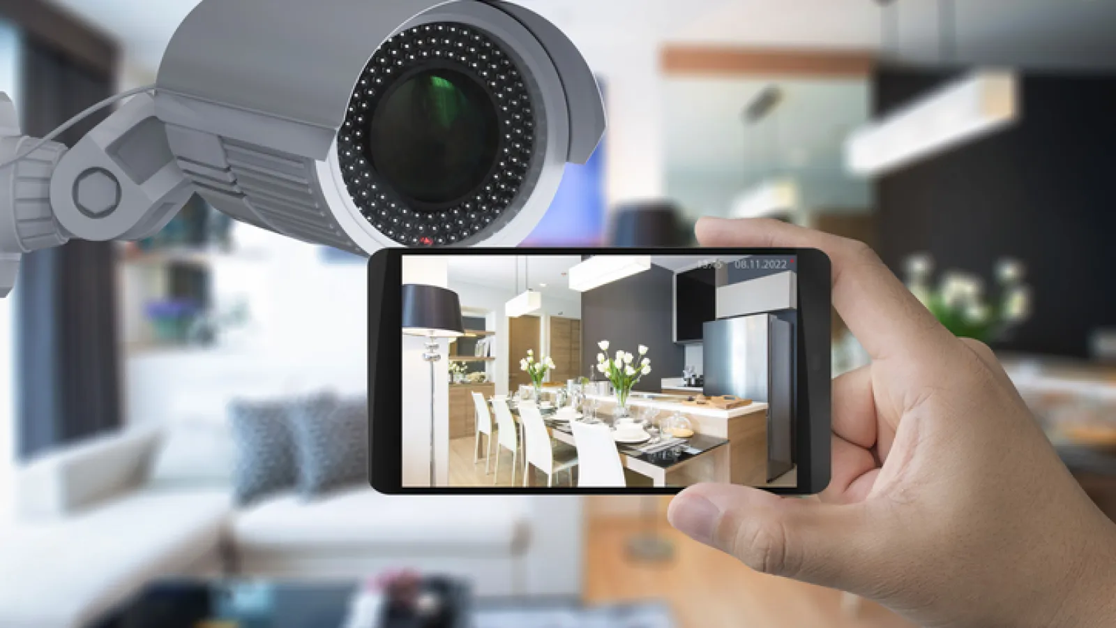 Hand holding smartphone showing security camera footage of modern dining area in a home interior environment.