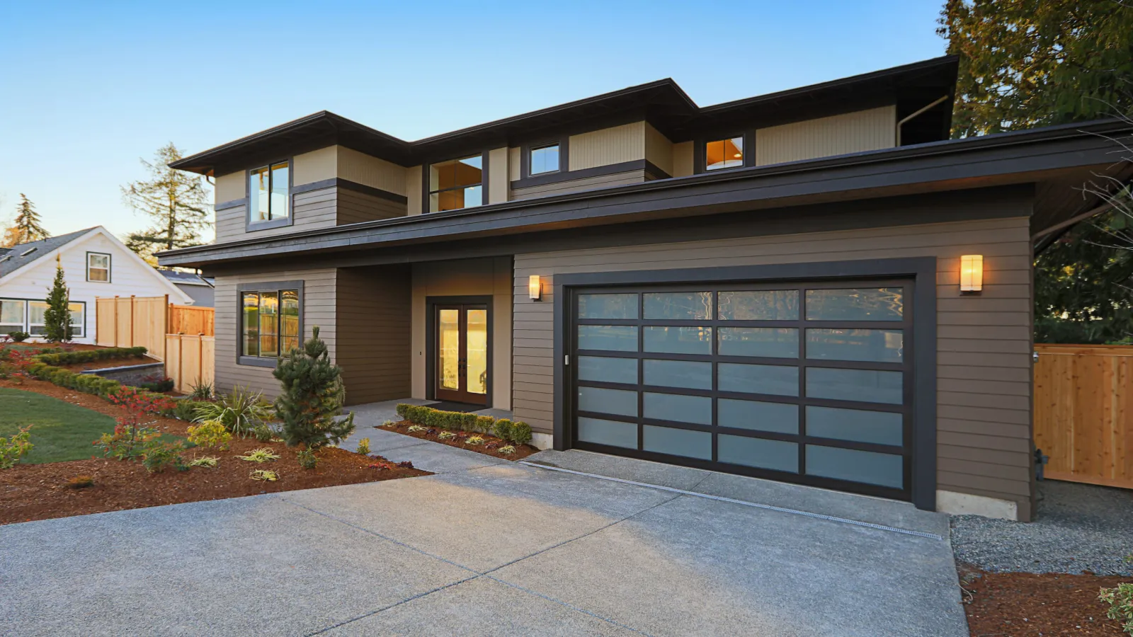 Modern two-story house with large glass garage door and twilight exterior lighting in suburban neighborhood.