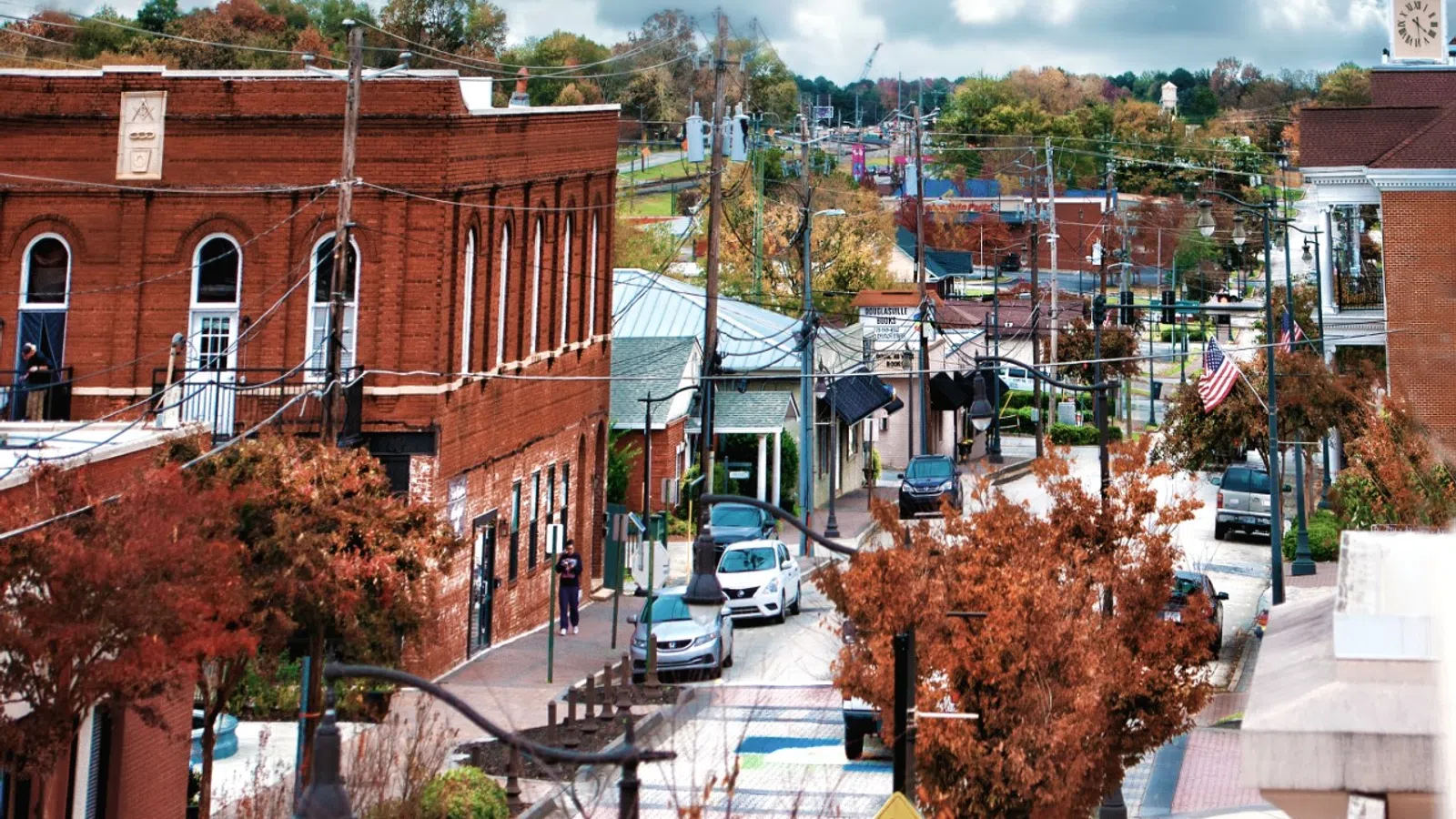 A scenic view of a downtown street lined with brick buildings, trees, and traffic under a cloudy sky.