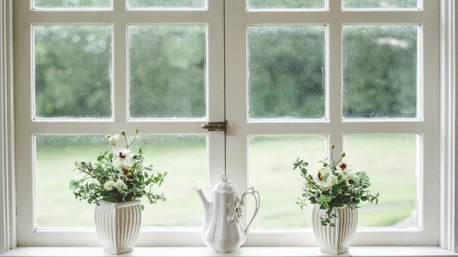 White ceramic teapot and two flower vases with greenery on a windowsill with bright natural light.
