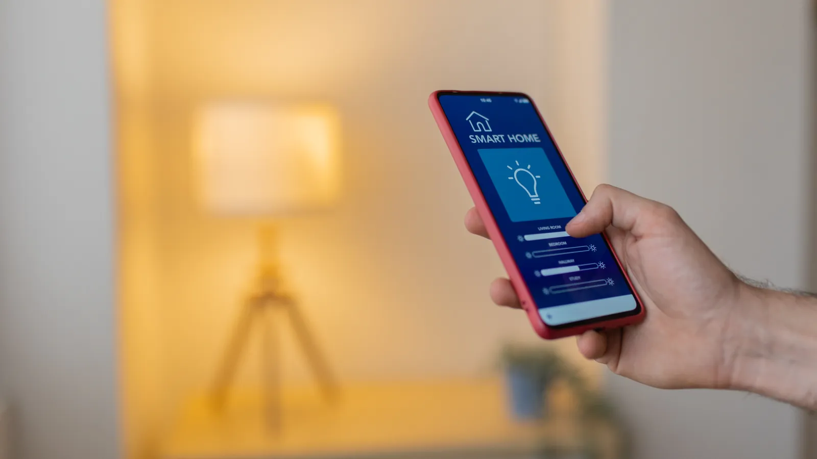Hand holding smartphone showing smart home app controlling lamp brightness in a cozy room.