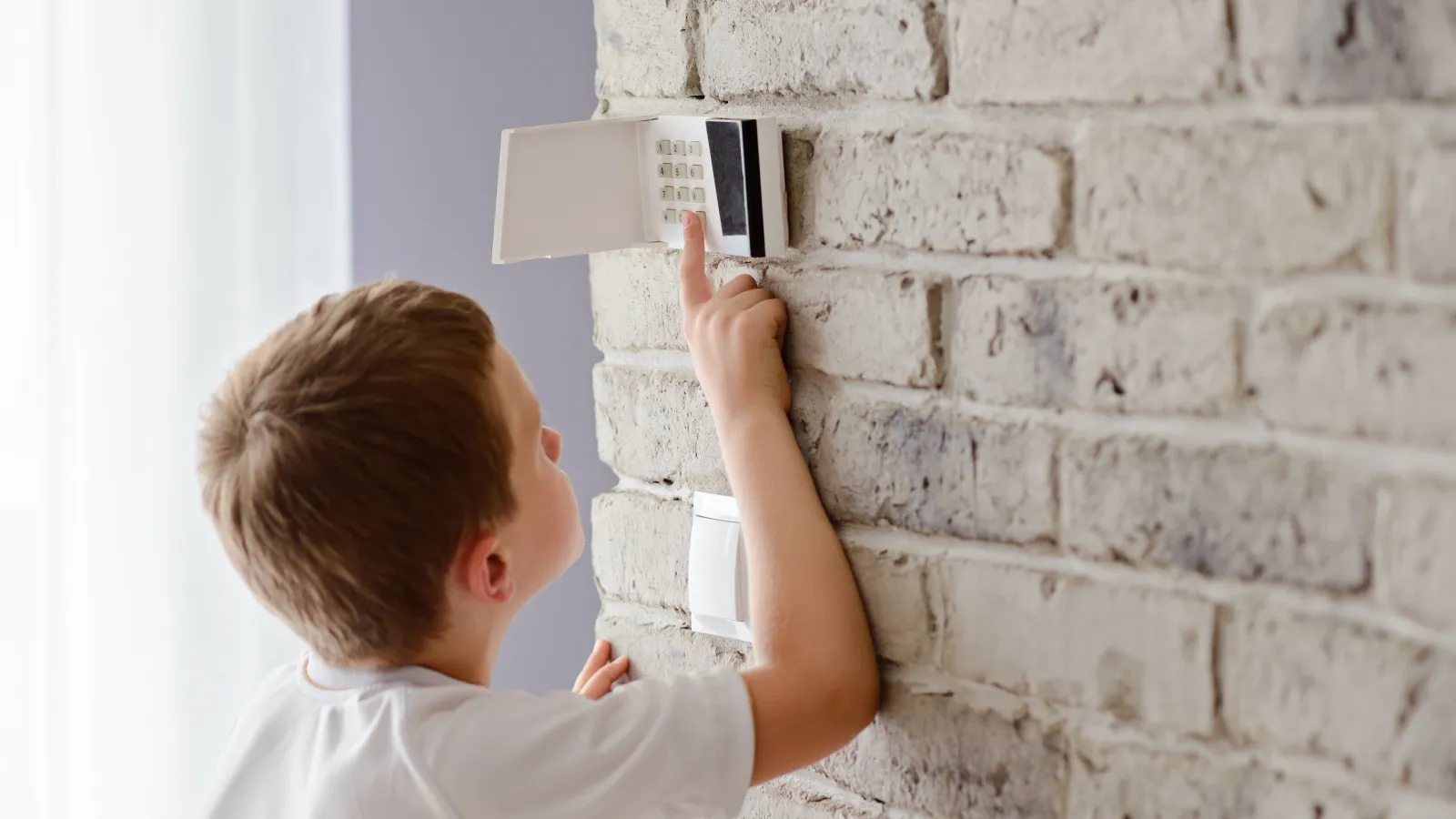 Young boy adjusting a white thermostat on a textured gray brick wall in a bright room.