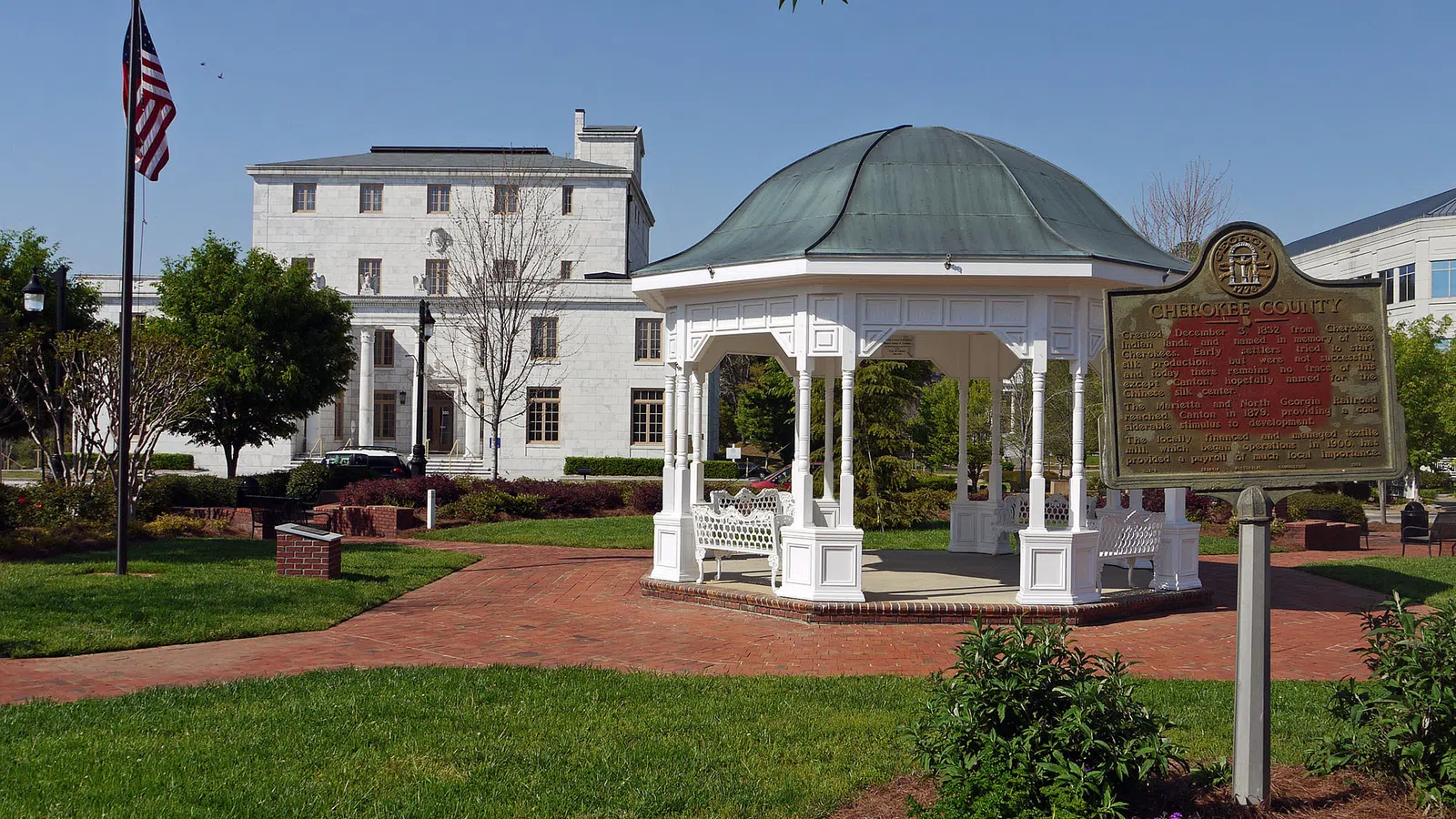 Scenic view of a gazebo in a park in Canton, GA with a historic marker and a flagpole under clear blue sky.