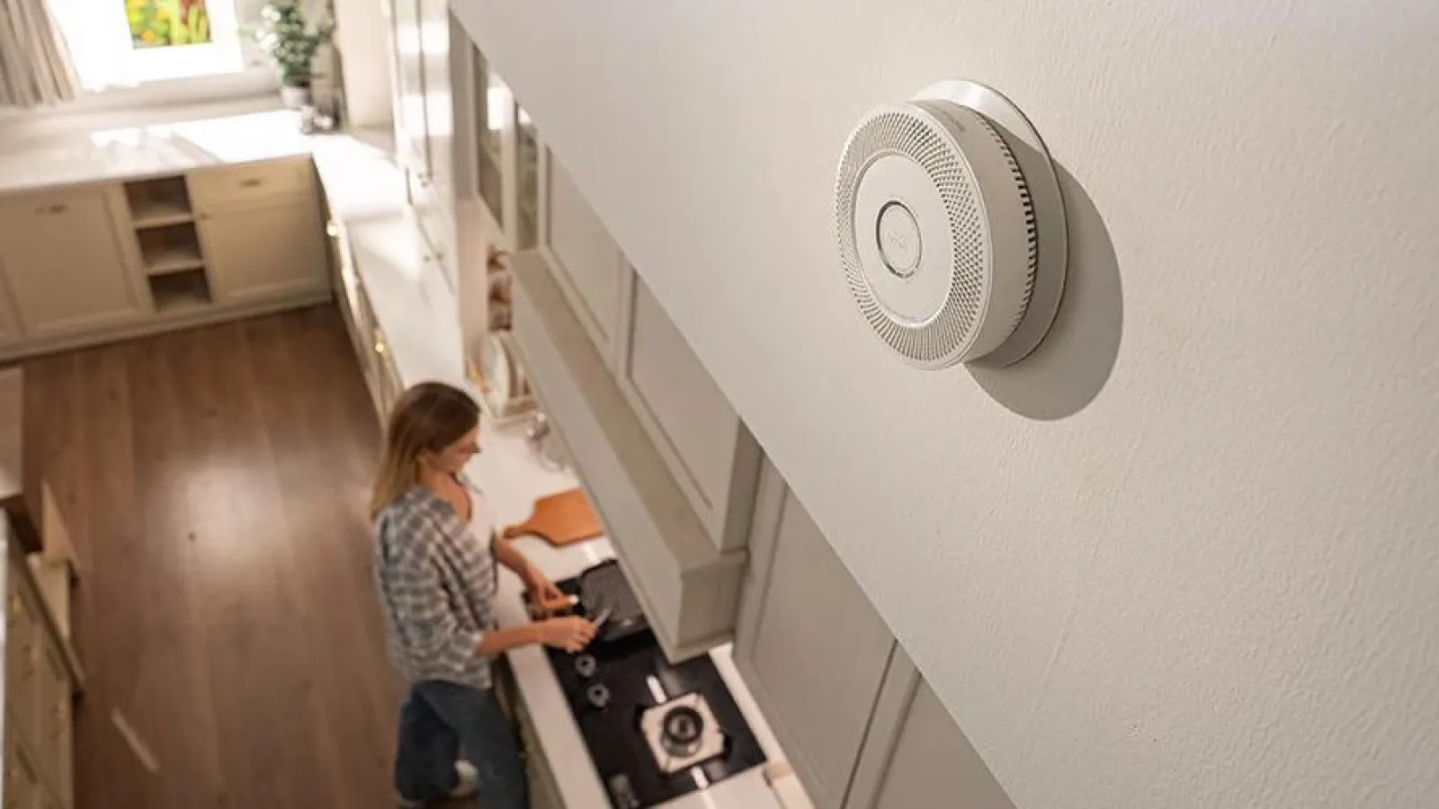 White smoke detector mounted on kitchen wall with woman cooking in modern kitchen background.