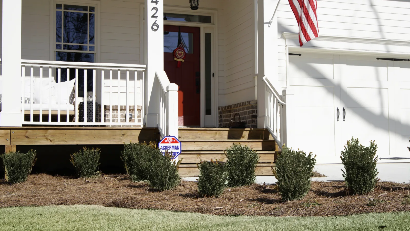 Front porch of a white house with American flag, red door, green bushes, and home security sign on lawn