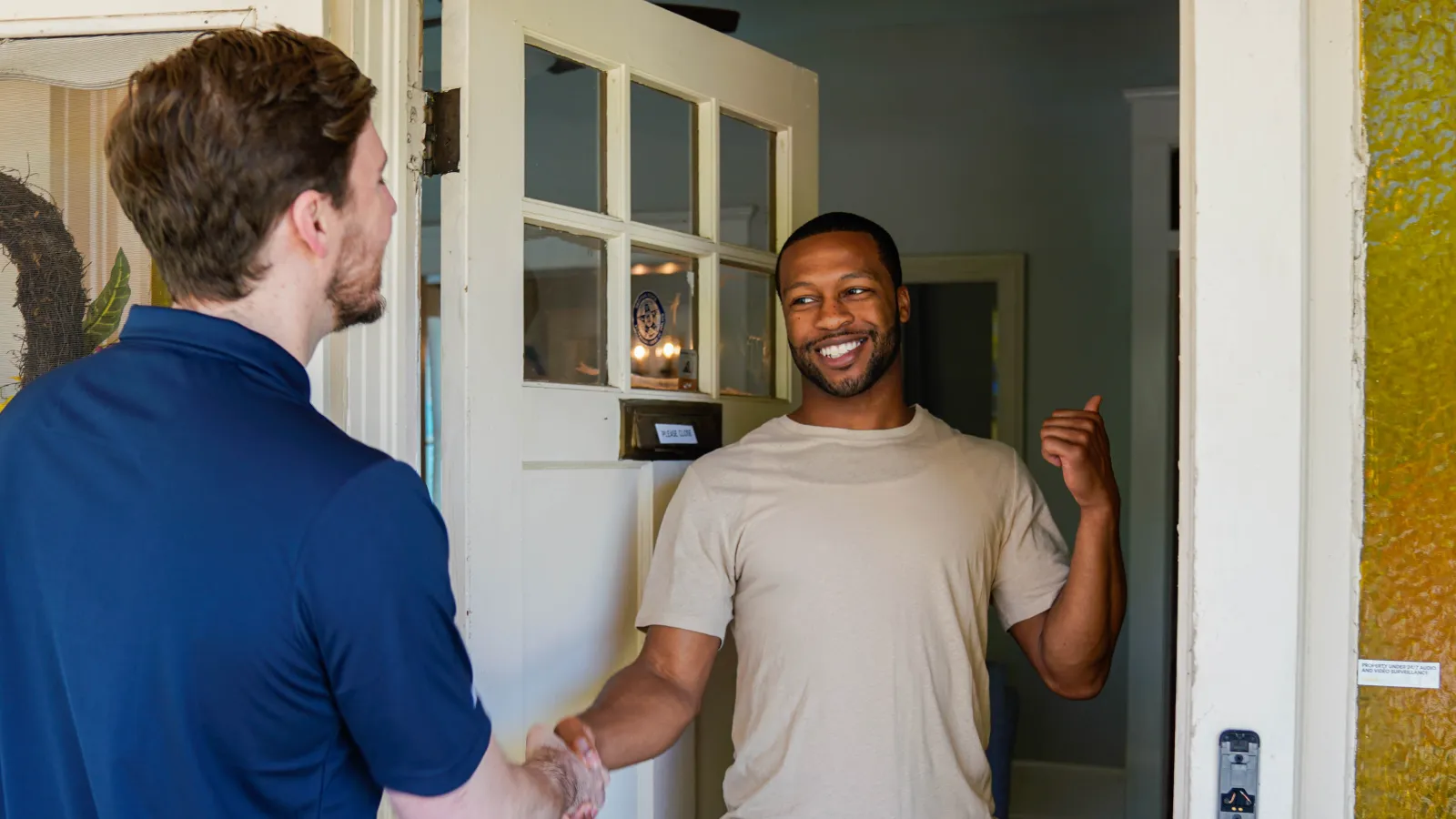Two men shaking hands at the doorway, one inviting the other inside with a smile in a home setting.