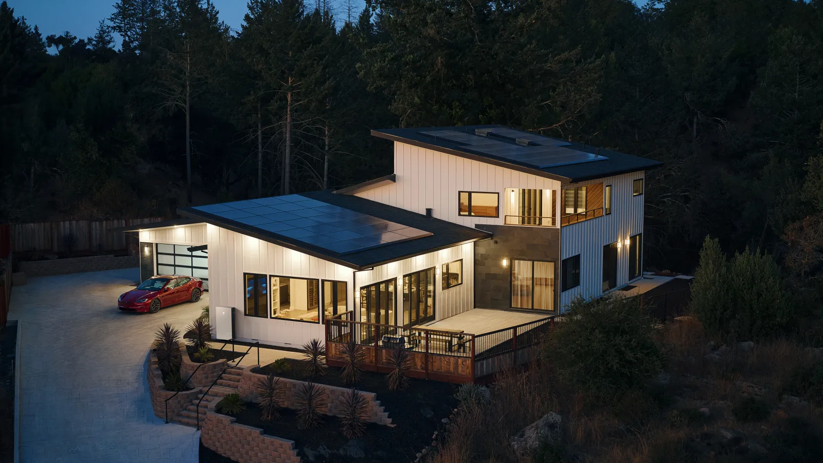 Modern white two-story house with solar panels and a red car in the driveway, surrounded by trees at dusk