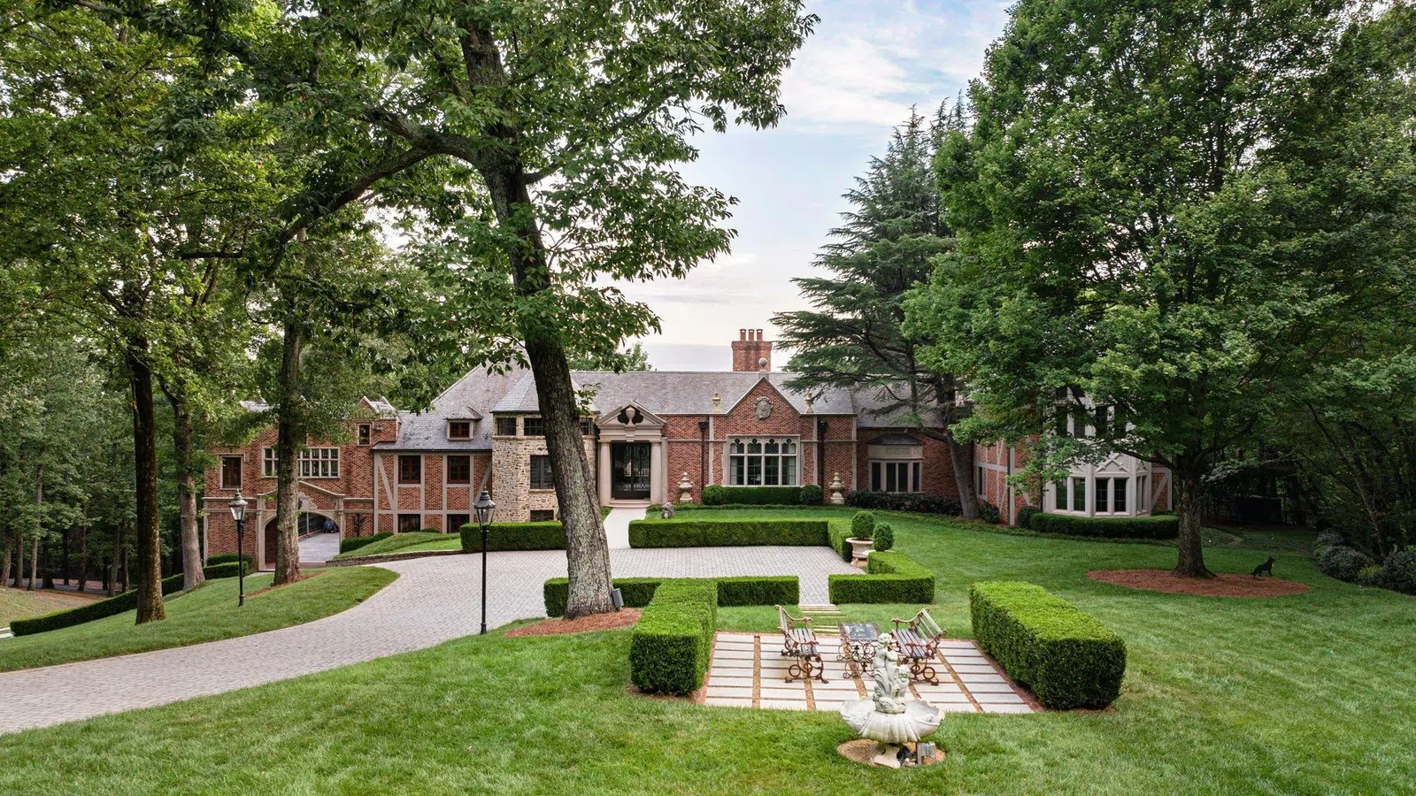 Large brick mansion with landscaped lawn, trimmed hedges, and outdoor seating area under tall trees.
