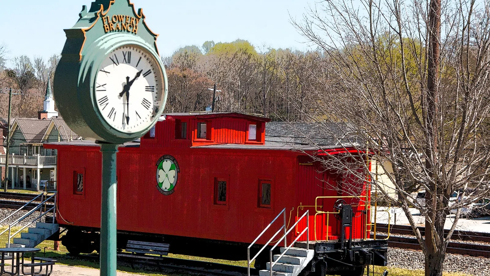 Historic red train car near a Flowery Branch clock in a small town park setting with bare trees and evergreen background.