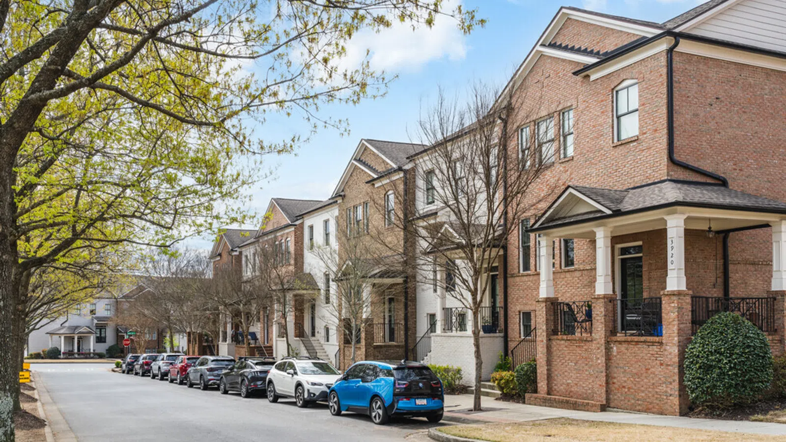 Row of modern brick and white townhouses with parked cars along a tree-lined suburban street on a clear day