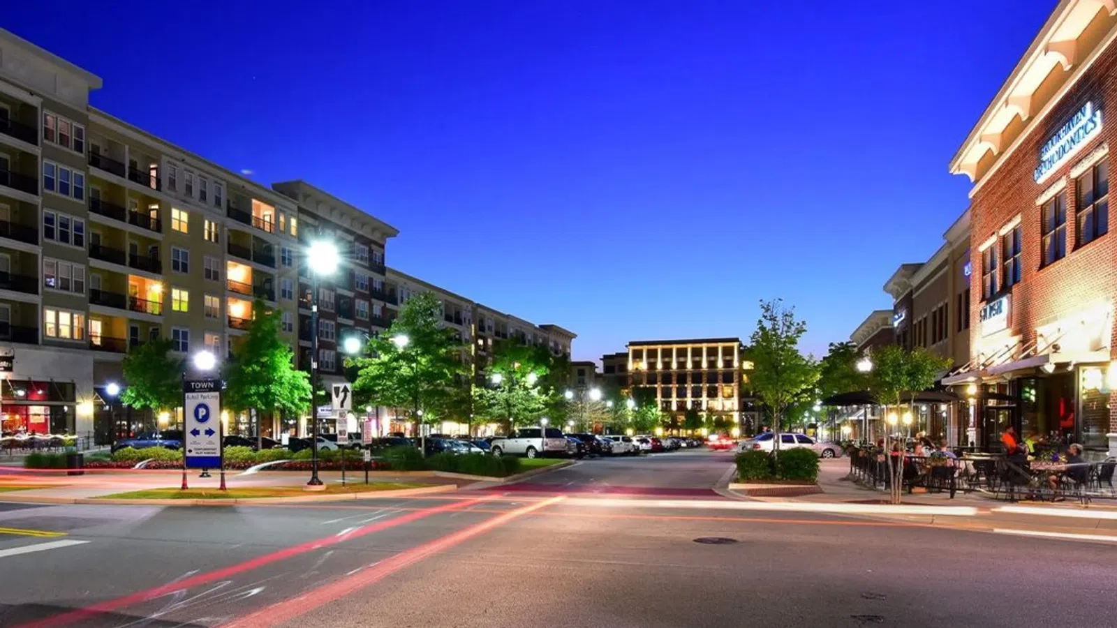 Night view of an urban street with modern apartments, lit trees, parked cars, and outdoor dining on the right.