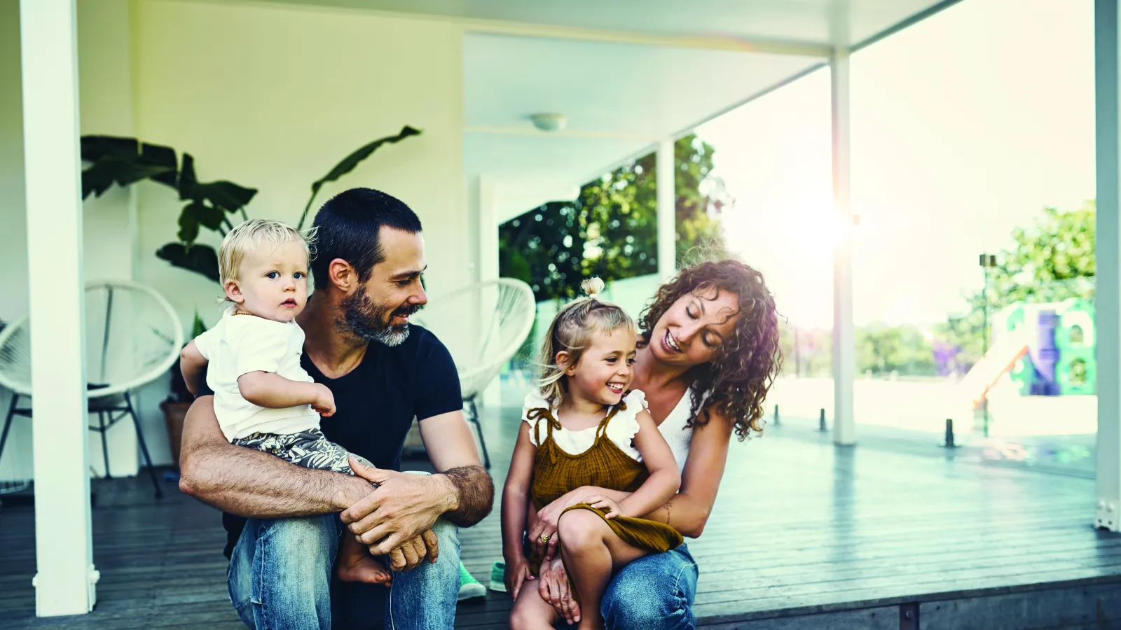 Happy family of four sitting on wooden porch steps in sunlight, enjoying quality time together outdoors.
