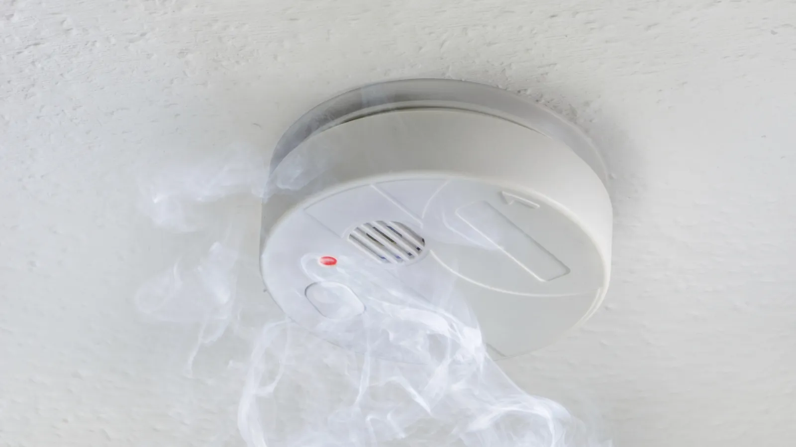 White smoke alarm mounted on ceiling with smoke rising indicating potential fire hazard.