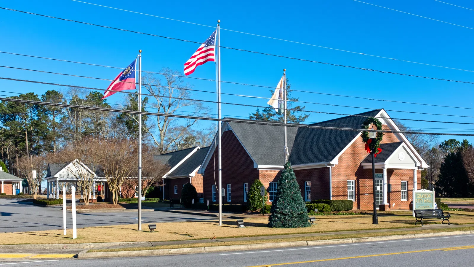 Brick town hall with American and state flags, wreath on lamp post, clear blue sky, and surrounding trees in small town.